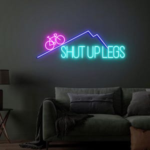 May include: A neon sign with a bicycle and mountain design in pink and blue. The words "SHUT UP LEGS" are written in bright turquoise. The sign is mounted on a dark wall above a green sofa.