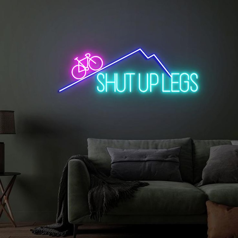 May include: A neon sign with a bicycle and mountain design in pink and blue. The words "SHUT UP LEGS" are written in bright turquoise. The sign is mounted on a dark wall above a green sofa.