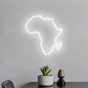 May include: A white neon light shaped like the African continent is fixed to a grey wall. Beneath, a small succulent plant in a white pot and a decorative wooden vase sit on a black surface.