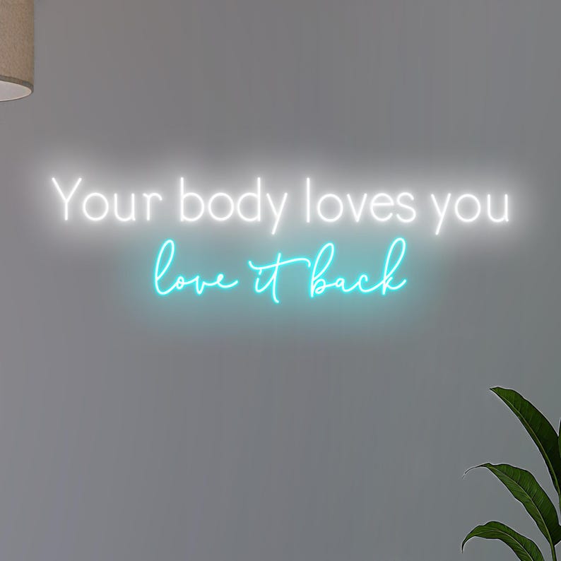 K&ouml;nnte beinhalten: Ein Neonschild mit dem Schriftzug "Your body loves you love it back" in Wei&szlig; und T&uuml;rkis. Das Schild ist an einer grauen Wand angebracht, mit einer Pflanze in der unteren rechten Ecke.