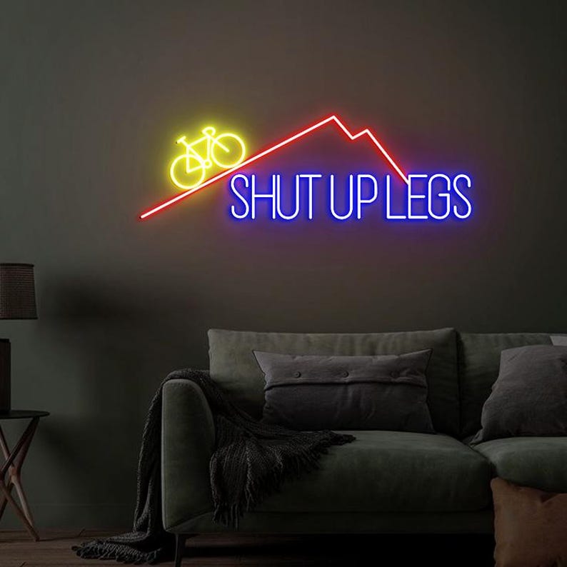 May include: Neon sign with a yellow bicycle, red mountain range, and the words "SHUT UP LEGS" in blue. The sign is mounted on a dark gray wall above a green sofa with pillows and a blanket.