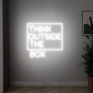 Może przedstawiać: Biały neon na szarej ścianie wyświetla frazę „THINK OUTSIDE THE BOX” w prostokątnej ramie. Znak jest podświetlony, tworząc jasną poświatę na neutralnym tle. W tle widoczne są oprawione dzieło sztuki i roślina doniczkowa.
