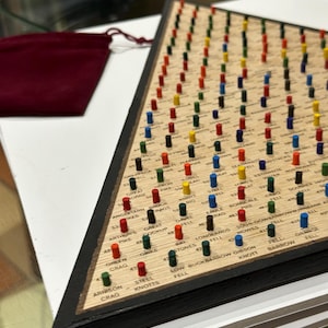May include: Triangular wooden board game with a black frame, featuring numerous coloured pegs. The board is labelled with place names and numbers. A small burgundy velvet bag is visible.
