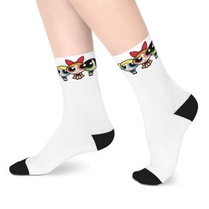 Può includere: Calzini bianchi con tallone e punta neri. I calzini presentano una grafica delle Powerpuff Girls,  Blossom, Bubbles e Buttercup, nelle loro pose caratteristiche.