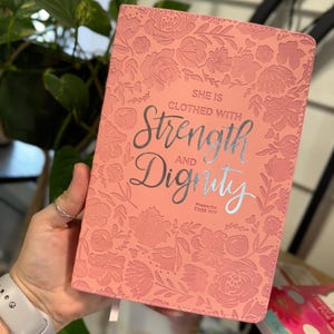 May include: A pink journal with a floral embossed design. The cover features the text "She is clothed with Strength and Dignity" and a Bible verse reference: Proverbs 31:25 NIV.