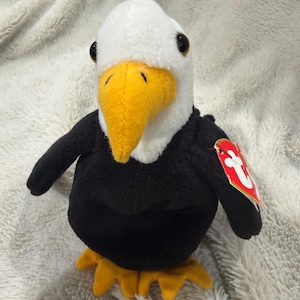 Könnte beinhalten: Ein Plüsch-Weißkopfseeadler mit weißem Kopf, leuchtend gelbem Schnabel und schwarzem Körper mit gelben Füßen. Das Stofftier hat dunkle, runde Augen und ein rotes, herzförmiges Etikett mit einem weißen T am linken Flügel befestigt.