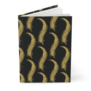 May include: A hardcover journal with a black cover featuring a repeating pattern of gold-colored feathers. The journal is open, revealing white lined pages. The spine and edges of the journal are also black.