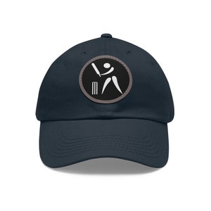 Puede incluir: Una gorra de béisbol azul marino con un gráfico de jugador de cricket blanco sobre un parche circular negro. El parche tiene un borde gris.