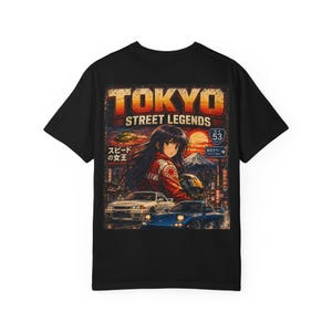 Peut inclure: T-shirt noir avec un motif graphique "TOKYO STREET LEGENDS". Le motif comprend une illustration de style anime d'une femme, des voitures et du texte japonais. Le design a un aspect vintage et vieilli.