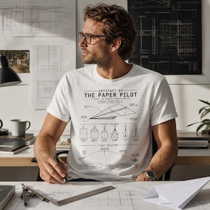 Peut inclure: T-shirt blanc avec un motif d'avion en papier et le texte "ARTIFACT 09 - THE PAPER PILOT". Le design comprend un schéma de la séquence de pliage et de la trajectoire de vol. Le t-shirt est porté par une personne.