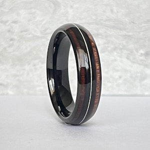 Könnte beinhalten: Ein schwarzer Ring mit Holzeinlage und silberfarbenem Akzent. Die Holzeinlage hat eine rotbraune Farbe. Der Ring liegt auf einer weißen Oberfläche.
