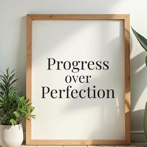 May include: A framed print with the words "Progress over Perfection" in black font. The frame is made of light-colored wood. A potted plant is visible on the left side of the frame.