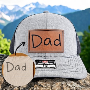 May include: A gray and black trucker hat with a brown leather patch that says "Dad" in handwritten font. The hat is adjustable and has a black mesh back.