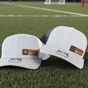 May include: Two gray and white baseball caps with leather patches that read "COACH Barrel" and "COACH David". One cap has a football graphic, the other a helmet. Both have a mesh back and a curved brim.