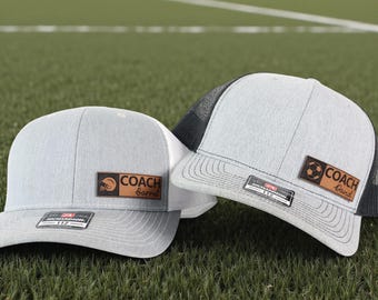 Peut inclure: Deux casquettes de baseball grises et blanches avec des écussons en cuir indiquant "COACH Barrel" et "COACH David". L'une présente un graphisme de football, l'autre un casque. Les deux ont un dos en filet et une visière incurvée.