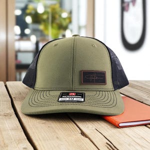 May include: An olive green trucker hat with a black mesh back. The hat features a rectangular brown patch with a signature design and a small tag that reads "RICHARDSON ORIGINAL 112 TRUCKER ADJUSTABLE".