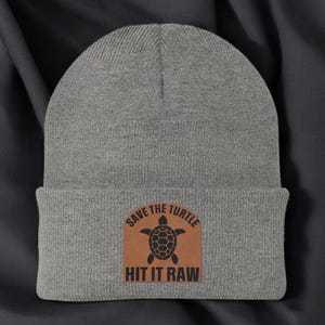 May include: A gray beanie with a brown patch featuring a black turtle graphic and the text "SAVE THE TURTLE" above and "HIT IT RAW" below. The beanie is made of a soft, knitted material.