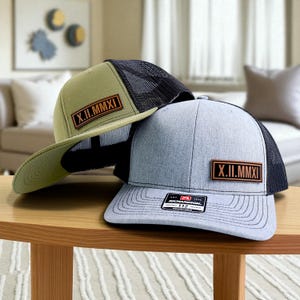 May include: Two baseball caps with mesh backs and leather patches. One cap is olive green with a black mesh back, the other is light gray with a black mesh back. Both have the Roman numeral "X.II.MMXI" on the leather patch.