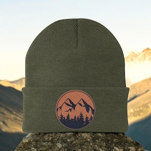 May include: Olive green beanie with a circular patch featuring a mountain range and pine trees in shades of brown and navy blue. The beanie is made of a soft, knitted material.
