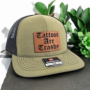 May include: A green and black trucker hat with a brown leather patch that says "Tattoos Are Trashy" in black lettering. The hat is adjustable and has a black mesh back.
