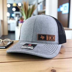 May include: Gray trucker hat with a black mesh back and a brown leather patch featuring the Texas state outline and the letters "TX." The hat has a curved brim and an adjustable strap. The hat is sitting on a wooden surface.