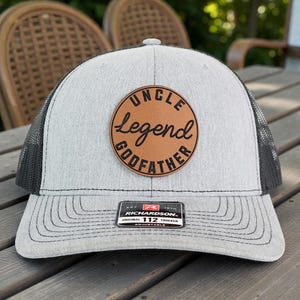 May include: Gray trucker hat with a black mesh back and a brown leather patch. The patch features the words "UNCLE LEGEND GODFATHER" in a circular design. The hat has a curved brim and an adjustable strap.