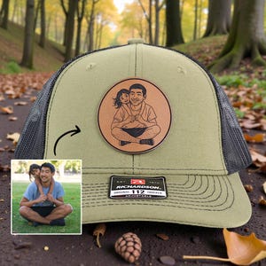 May include: Olive green trucker hat with a black mesh back and a leather patch. The patch features a sketch of a person and a child. The hat has a Richardson 112 tag.