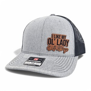 Peut inclure: Un chapeau de camionneur gris et noir avec un patch en cuir brun qui dit "J'aime ma vieille dame un peu plus jeune".