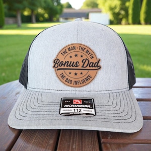 May include: A gray and black trucker hat with the text "Bonus Dad" on a brown leather patch. The hat has a curved brim and an adjustable strap. The patch also reads "The Man, The Myth, The Bad Influence."