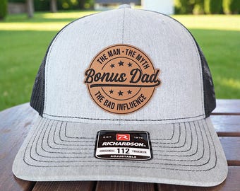 May include: A gray and black trucker hat with the text "Bonus Dad" on a brown leather patch. The hat has a curved brim and an adjustable strap. The patch also reads "The Man, The Myth, The Bad Influence."