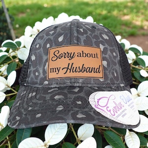 May include: A black trucker hat with a leopard print design. The hat features a brown leather patch with the words "Sorry about my Husband" in black script. The hat has a mesh back and a curved brim.