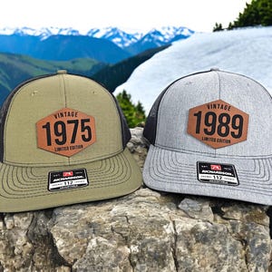 May include: Two vintage trucker hats with black mesh back panels. The hats have a brown leather patch with the text "Vintage" and a year, "1975" on one hat and "1989" on the other. The text "Limited Edition" is printed below the year on each patch.