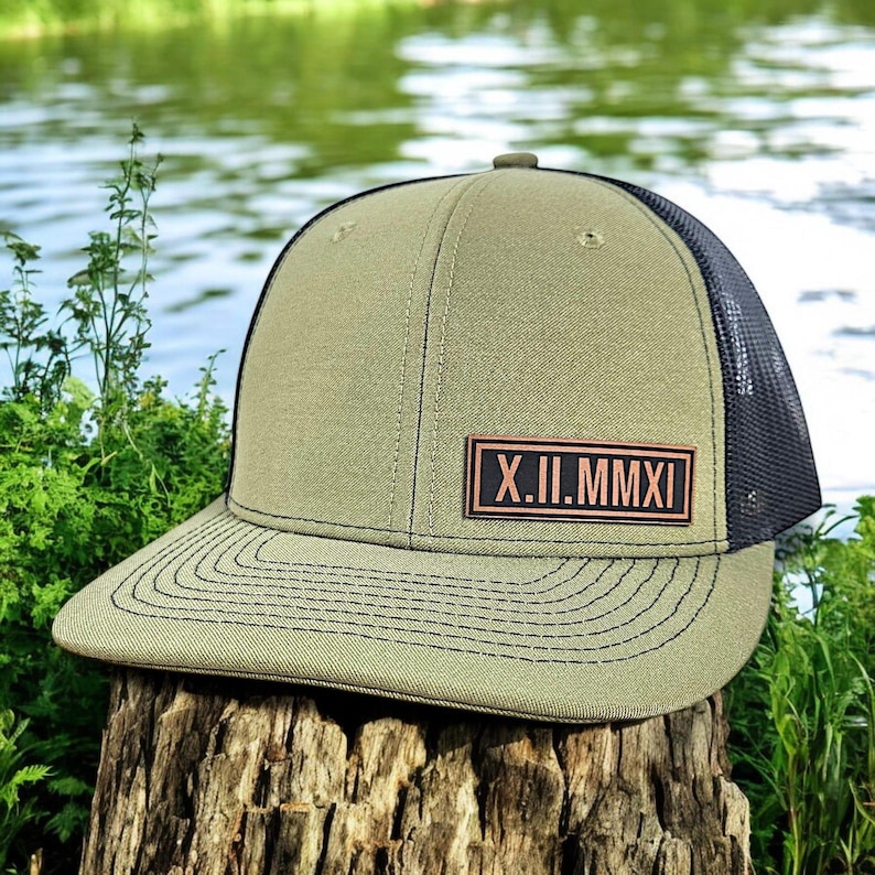 May include: Olive green trucker hat with a black mesh back and a curved brim. The hat features a leather patch with the Roman numerals X.II.MMXI. The hat is sitting on a tree stump with a blurred water background.