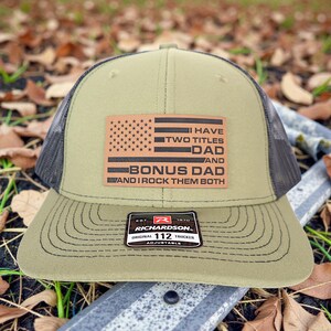 May include: Olive green trucker hat with a black mesh back and a brown leather patch featuring an American flag design and the text "I HAVE TWO TITLES DAD AND BONUS DAD AND I ROCK THEM BOTH". The hat has a curved brim and an adjustable strap.