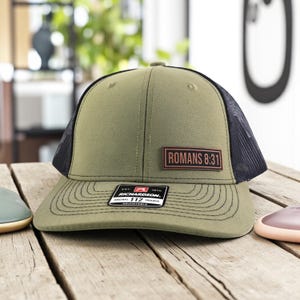 May include: Olive green trucker hat with a black mesh back and a curved brim. The hat features a rectangular patch with the text "ROMANS 8:31". A small tag on the brim reads "RICHARDSON 112 TRUCKER".