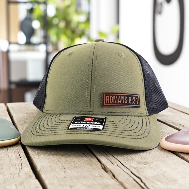 May include: Olive green trucker hat with a black mesh back and a curved brim. The hat features a rectangular patch with the text "ROMANS 8:31". A small tag on the brim reads "RICHARDSON 112 TRUCKER".
