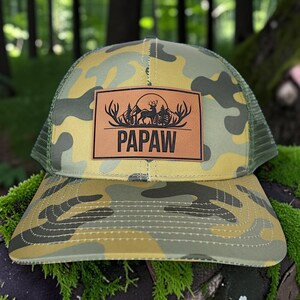 May include: A camouflage trucker hat with a brown leather patch. The patch features a deer silhouette, trees, and the word "PAPAW" in bold letters. The hat has a green mesh back and a curved brim.