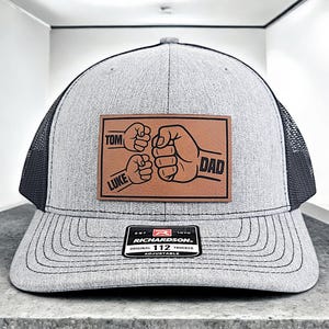 May include: A gray and black trucker hat with a brown leather patch that says "TOM, LUKE, DAD" with a fist bump graphic. The hat has a black mesh back and an adjustable strap with the Richardson 112 logo.