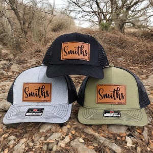 May include: Three trucker hats in black, gray, and olive green. Each hat features a brown leather patch with the name "Smiths" and the word "Family" underneath. The hats have a mesh back and a curved brim.
