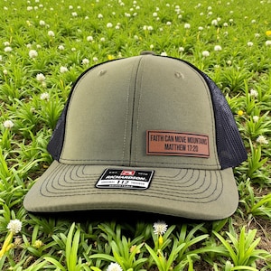 Puede incluir: Gorra de camionero verde oliva con parte trasera de malla negra y visera curva. Un parche rectangular en la parte delantera dice "FAITH CAN MOVE MOUNTAINS MATTHEW 17:20". La gorra tiene una etiqueta Richardson 112.
