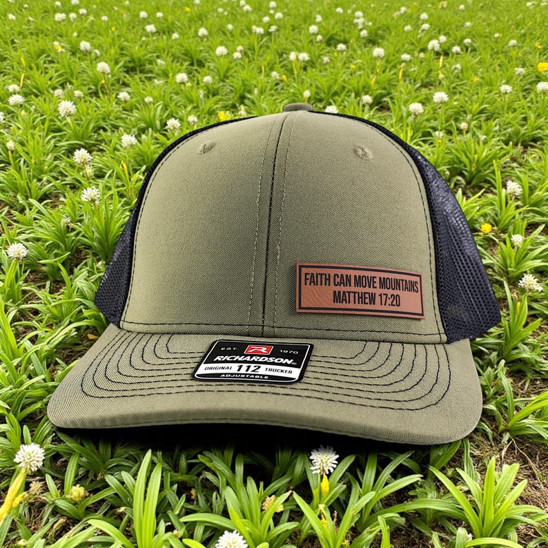 Puede incluir: Gorra de camionero verde oliva con parte trasera de malla negra y visera curva. Un parche rectangular en la parte delantera dice "FAITH CAN MOVE MOUNTAINS MATTHEW 17:20". La gorra tiene una etiqueta Richardson 112.