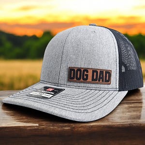May include: A gray and black trucker hat with a brown leather patch that says "DOG DAD".