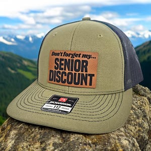 May include: A green and black trucker hat with a brown leather patch that says "Don't forget my... SENIOR DISCOUNT". The hat is adjustable and has a Richardson 112 logo.