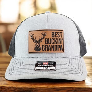 May include: A gray and black trucker hat with a leather patch that reads "BEST BUCKIN' GRANDPA" with a deer graphic. The hat has a curved brim and an adjustable strap. The Richardson brand label is visible.