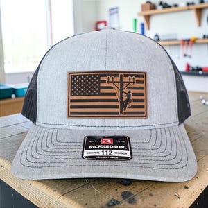 May include: A gray trucker hat with a black mesh back and a curved brim. The front features a brown leather patch with an American flag design and a silhouette of a lineman on a utility pole. The hat has a Richardson patch.