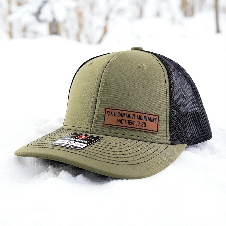 Puede incluir: Gorra de camionero verde oliva y negra con visera curva. Cuenta con un parche de cuero marr&oacute;n con el texto "FAITH CAN MOVE MOUNTAINS MATTHEW 17:20". La gorra est&aacute; en la nieve.