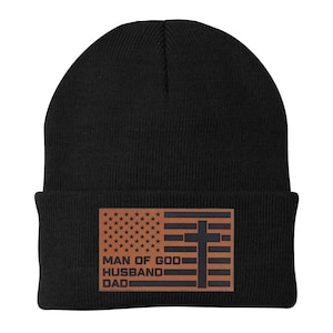 May include: Black beanie with a brown leather patch featuring an American flag design and a cross. The patch includes the text "MAN OF GOD, HUSBAND, DAD". The beanie is made of a soft, knitted material.