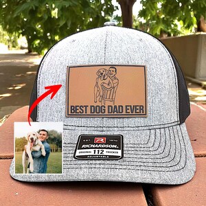 May include: Gray and black trucker hat with a leather patch that reads "BEST DOG DAD EVER." The patch features a graphic of a person holding a dog. The hat also has a Richardson patch.