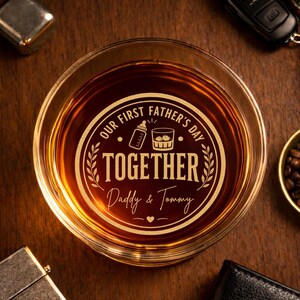 May include: A clear glass tumbler filled with amber liquid, featuring etched text that reads "OUR FIRST FATHER'S DAY TOGETHER Daddy & Tommy." The design includes a baby bottle, a whiskey glass, and a heart.