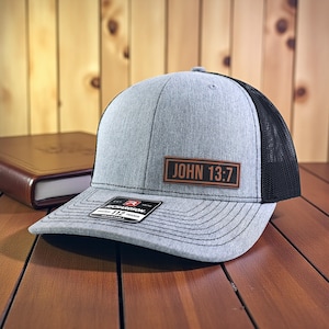 May include: A gray trucker hat with a black mesh back. The hat features a brown rectangular patch with the text "JOHN 13:7". The hat is sitting on a wooden surface.